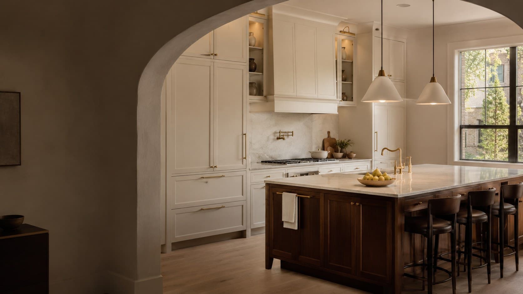 Cream painted kitchen seen through a plaster arch, with a walnut island, leather counter stools and conical pendants