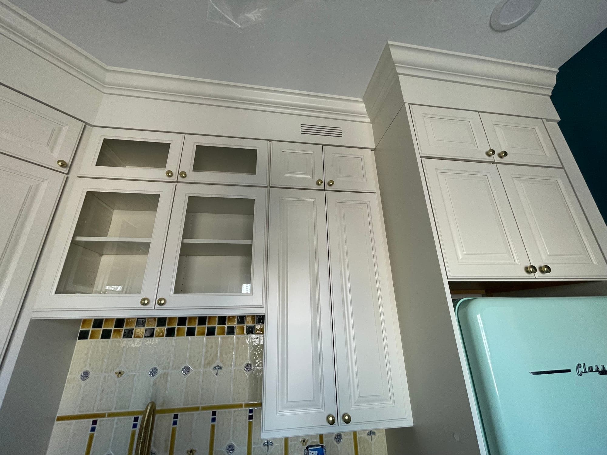 Cream raised panel cabinets with glass fronts and brass knobs under crown molding, beside a mint refrigerator