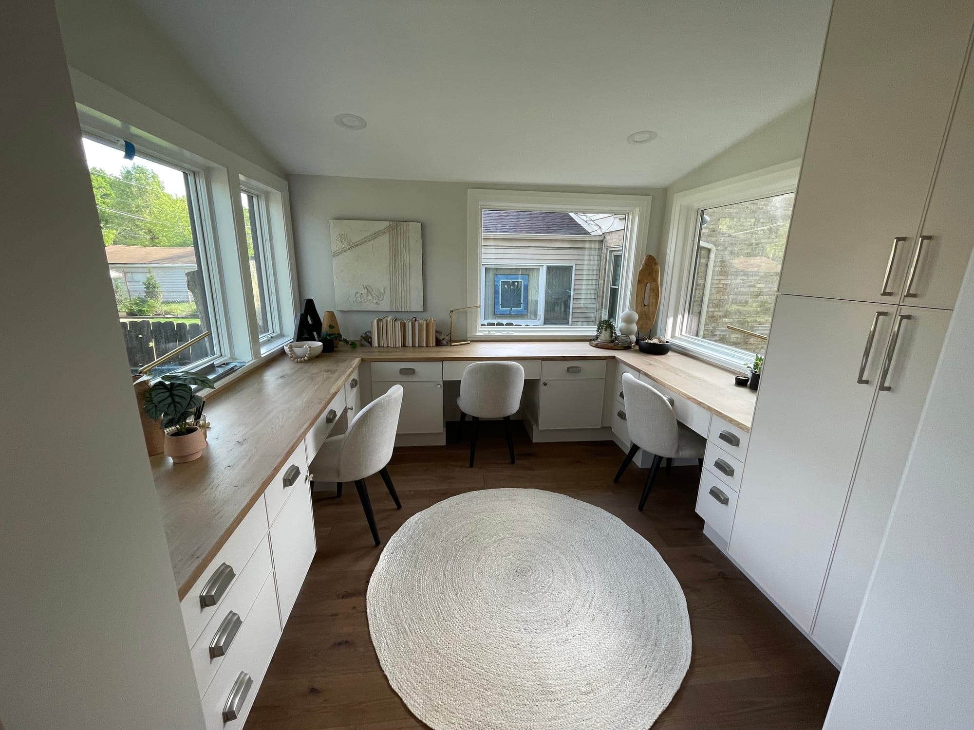 Home office with white built-in desk cabinets, white oak counters under three walls of windows and tall storage