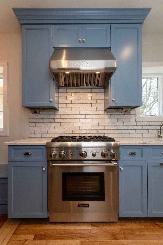 Blue gray cabinets with crown molding around a stainless hood and range, white brick-shaped tile and veined counters