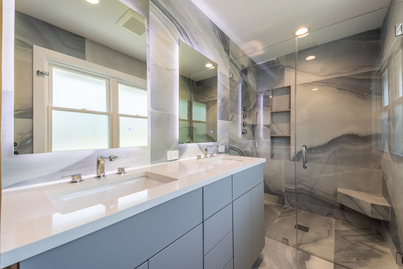 Bathroom with onyx look tile, a gray double vanity, backlit mirrors and a glass shower with a bench