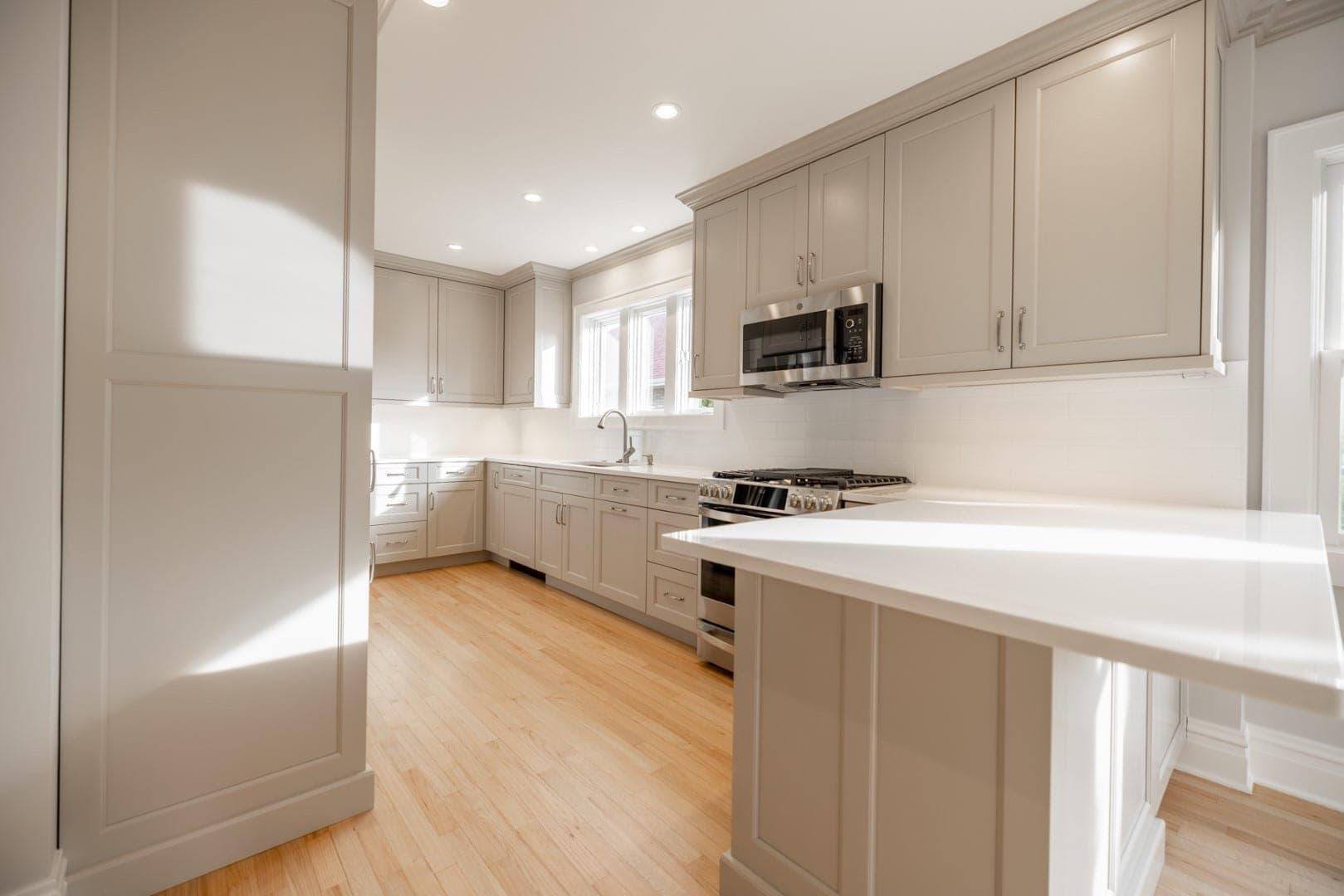 Sunlit U-shaped kitchen with greige Shaker cabinets, white counters, white tile and light wood floors
