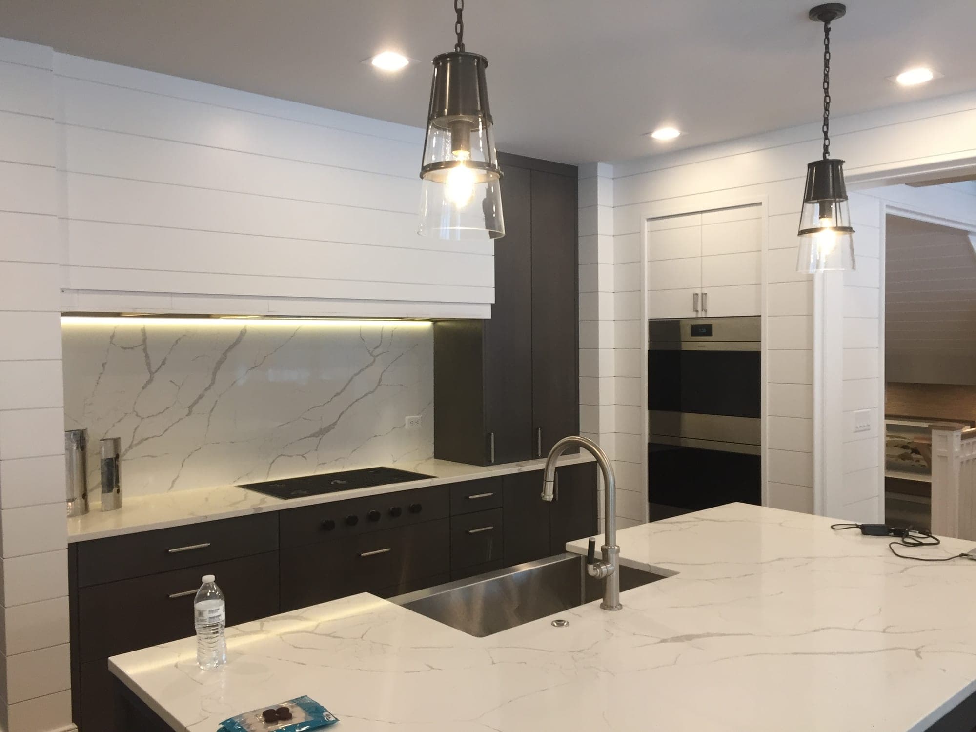 Cocina con isla veteada y tarja de acero inoxidable, lámparas colgantes de vidrio, gabinetes oscuros y hornos de pared