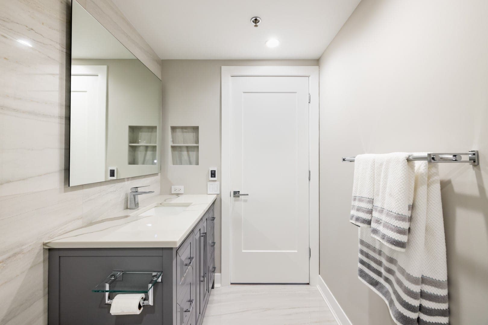 Mueble de lavabo gris con cubierta blanca contra un muro de losa veteada, junto a un muro pintado con repisa empotrada