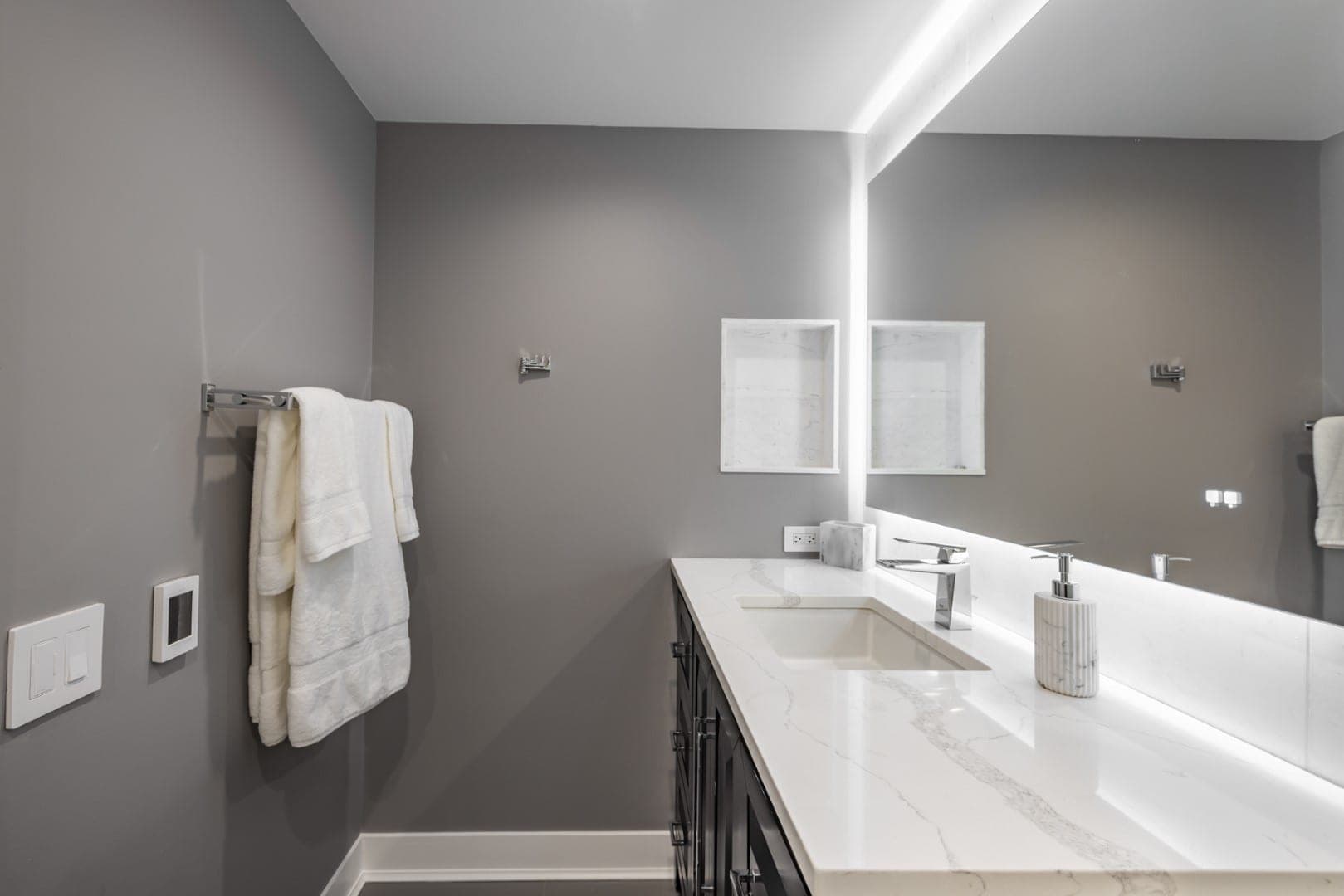 Baño gris con mueble de lavabo largo, cubierta blanca veteada, espejo iluminado en los bordes y repisa empotrada