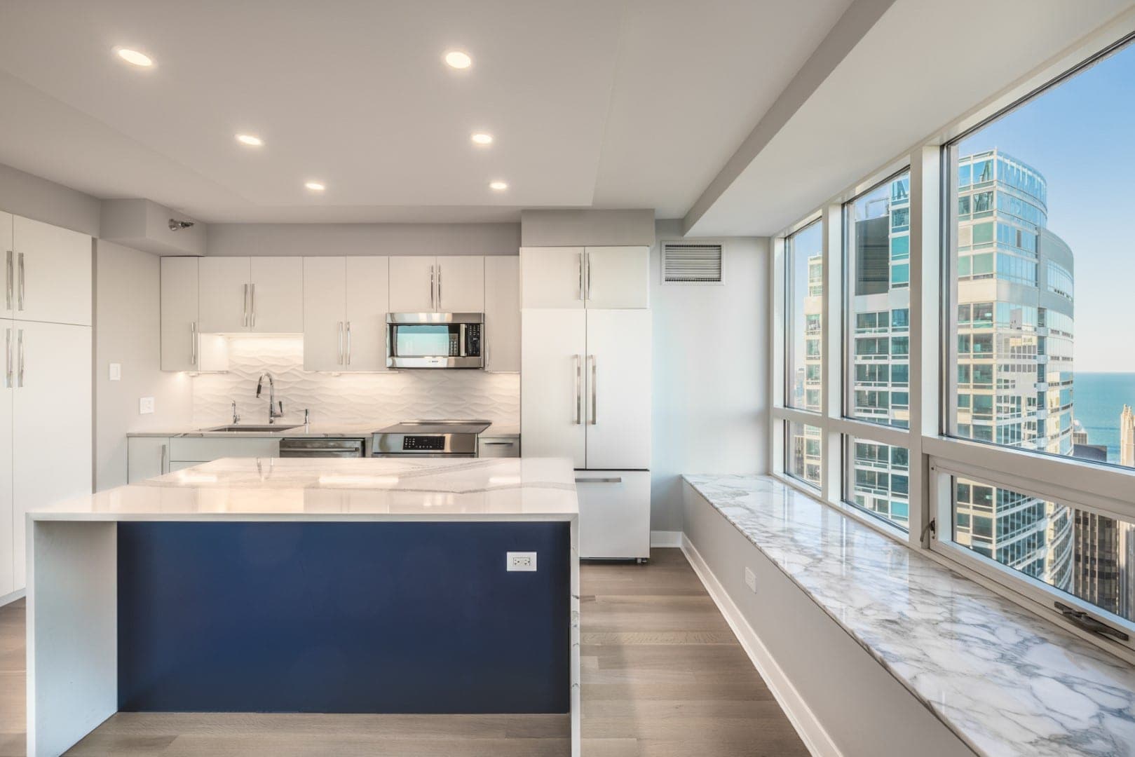 Cocina de condominio con isla azul marino bajo cubierta blanca veteada y repisa veteada a lo largo de ventanales de piso a techo
