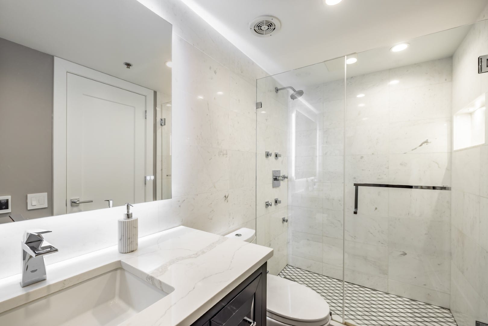 Baño con regadera walk-in de vidrio en azulejo blanco veteado con chorros corporales y nicho, junto a un espejo iluminado