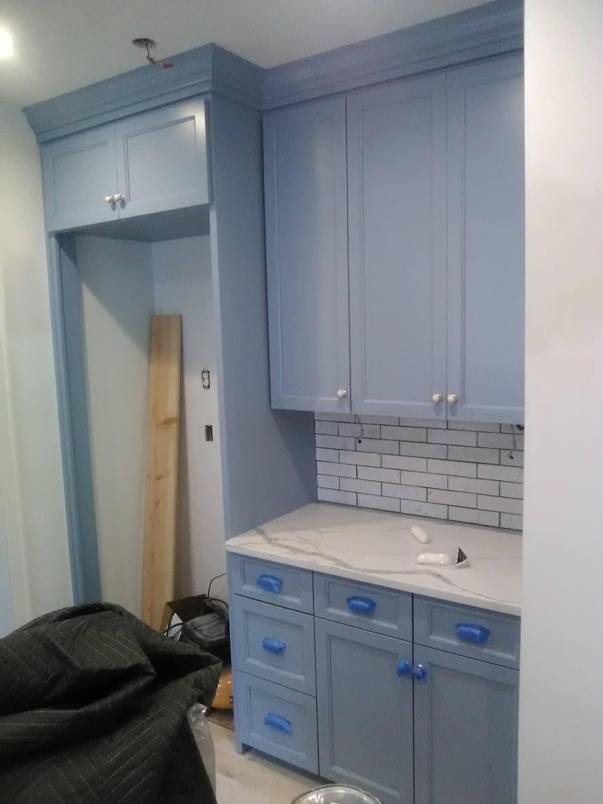 Blue-grey inset cabinets, a tall refrigerator surround and crown moulding installed, with the pulls still taped and a veined quartz counter set