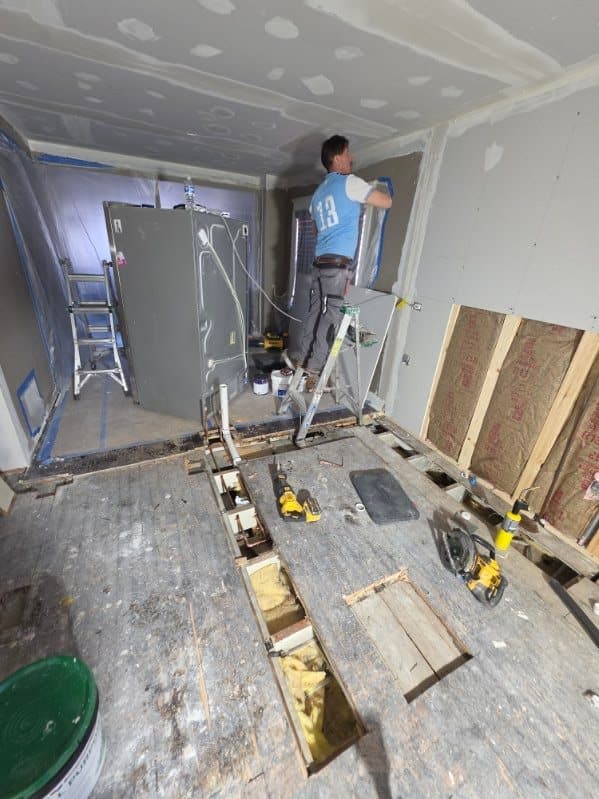 New drywall on the walls and ceiling while new drain lines are set into channels cut in the subfloor, with insulation in an open wall