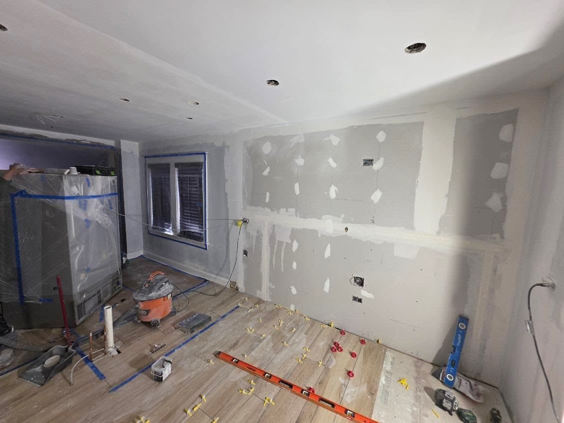 Taped and mudded drywall with outlet boxes marked, and wood-look floor tile being set with leveling clips and a long level