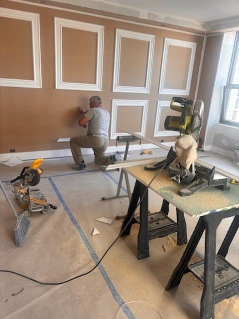 Carpenter kneeling at a wall to fit panel moulding, with two miter saws and a work table on the protected floor