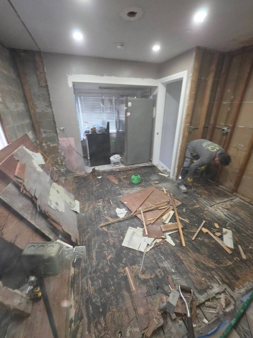 Kitchen taken back to the studs and the old subfloor, with torn-out lath and flooring on the floor and the refrigerator moved to the doorway
