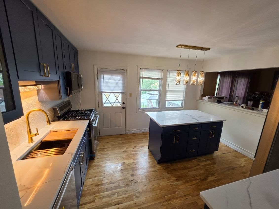 The finished kitchen with navy Shaker cabinets, brass pulls and faucet, a white island with seating, a herringbone tile backsplash and oak floors