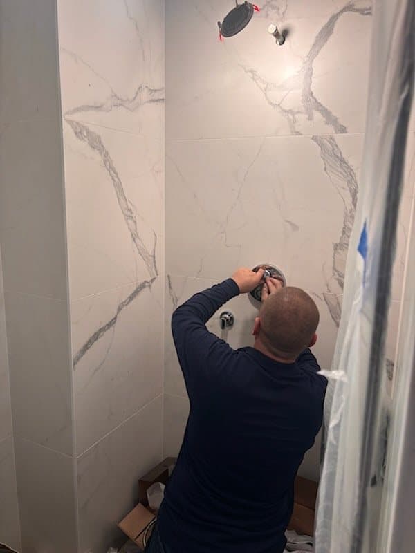 Plumber fitting the shower valve trim on a wall of large-format marble-look porcelain, with the ceiling shower head already in