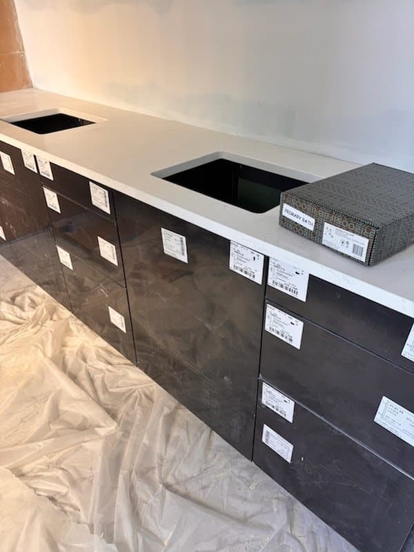 Long dark vanity with the labels still on the drawers and a white quartz top with two sink cutouts, before the sinks and faucets go in