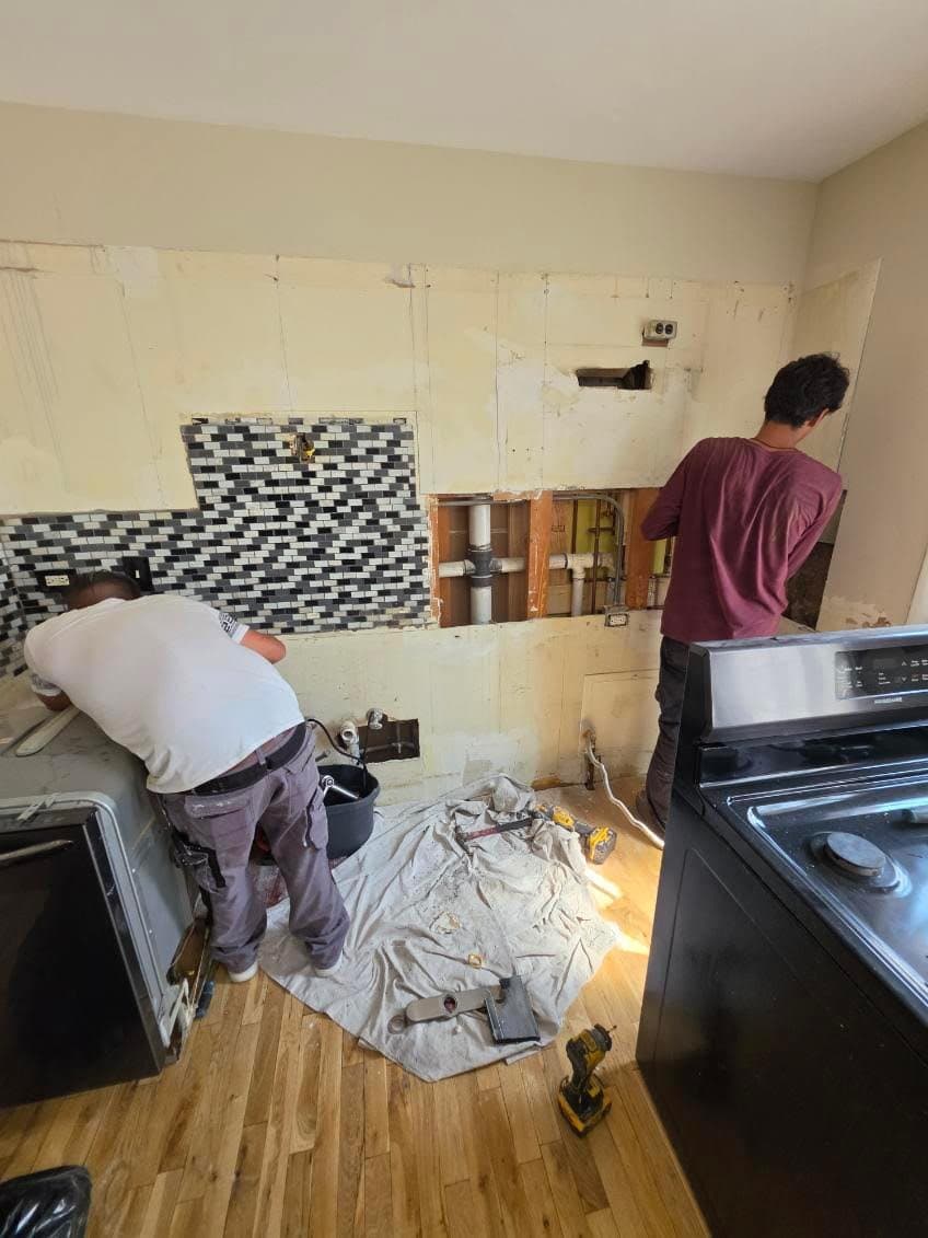 Two workers at a kitchen wall opened to the studs, with the old mosaic backsplash half removed and new supply and drain lines exposed
