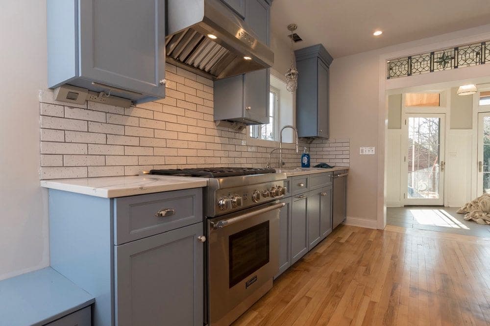 Cocina con gabinetes shaker gris azulado, estufa y campana de acero inoxidable, azulejo blanco tipo ladrillo y pisos de madera clara