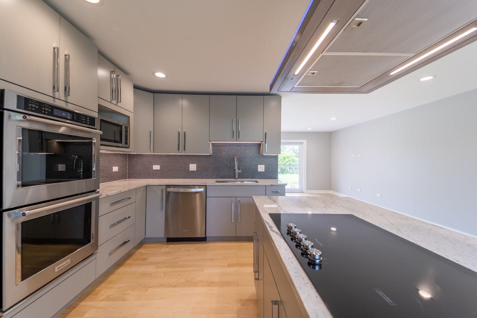 Kitchen with gray slab cabinets, dark penny tile, double wall ovens and an island cooktop under a ceiling hood