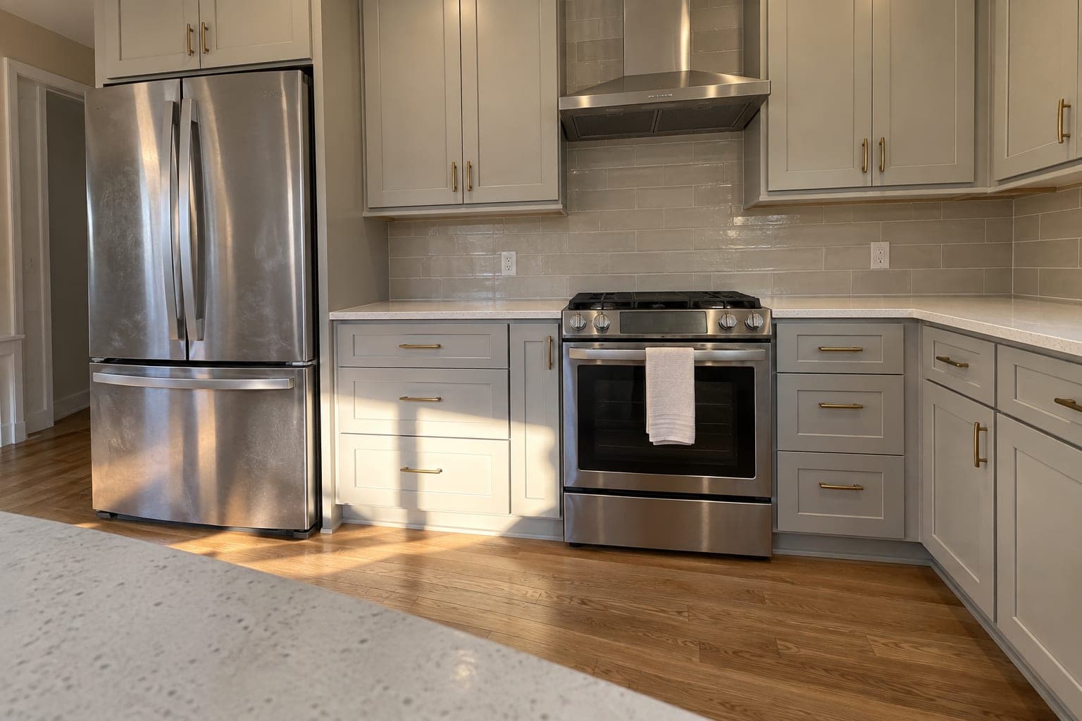 Appliance wall with warm gray Shaker cabinets, brass pulls, quartz counters, glossy greige tile, a stainless refrigerator, a range and a chimney hood