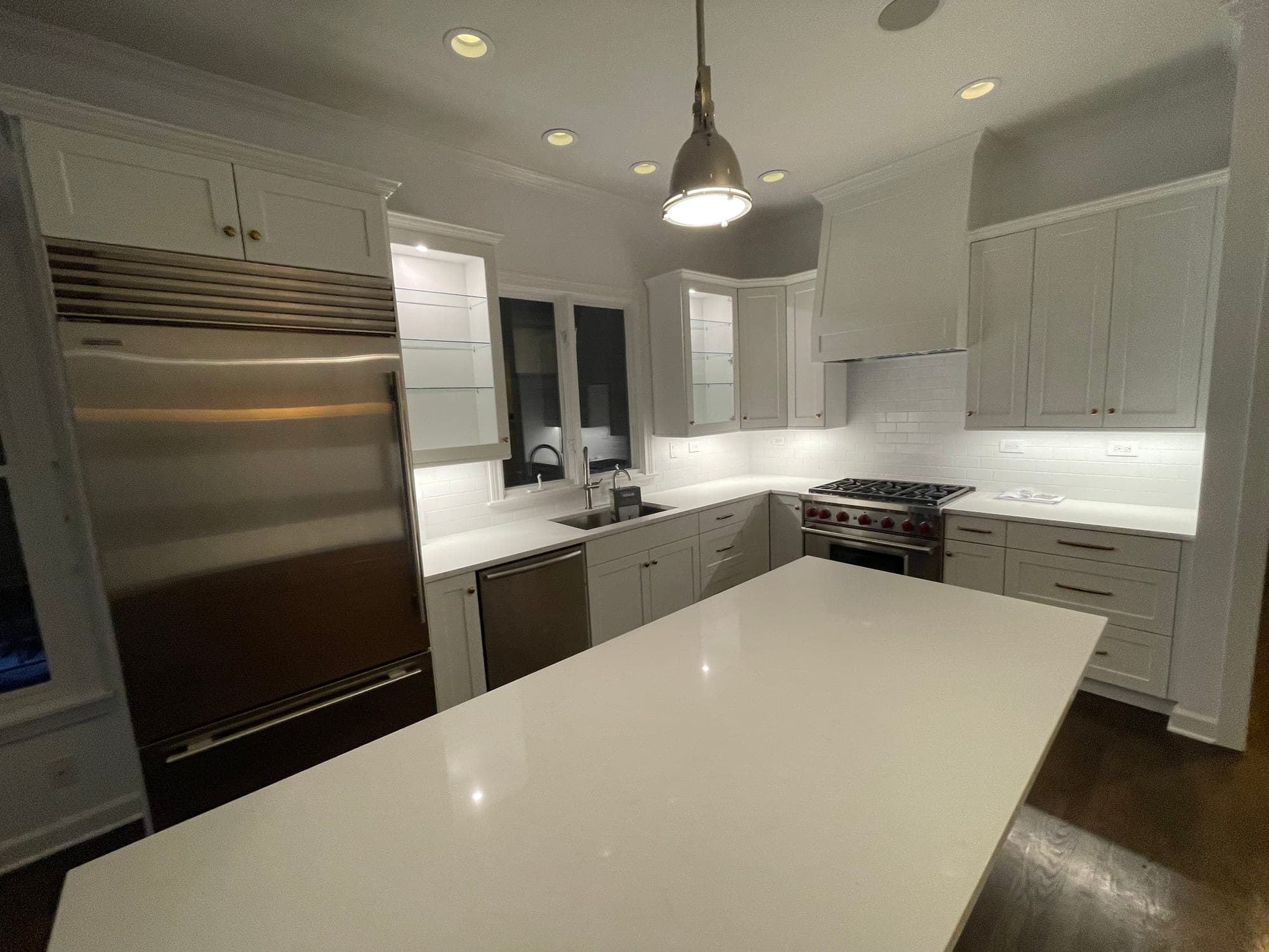 White Shaker kitchen at night with lit glass cabinets, a stainless refrigerator, a red-knob range and a white island