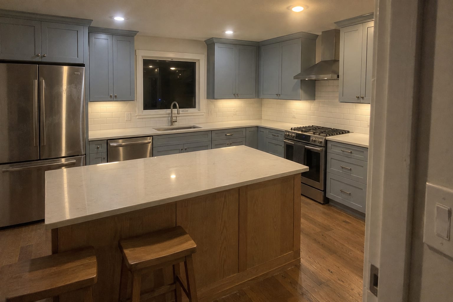 Kitchen with blue gray Shaker perimeter cabinets, a natural oak island with a quartz top and two stools, white subway tile, stainless appliances and a refinished oak floor