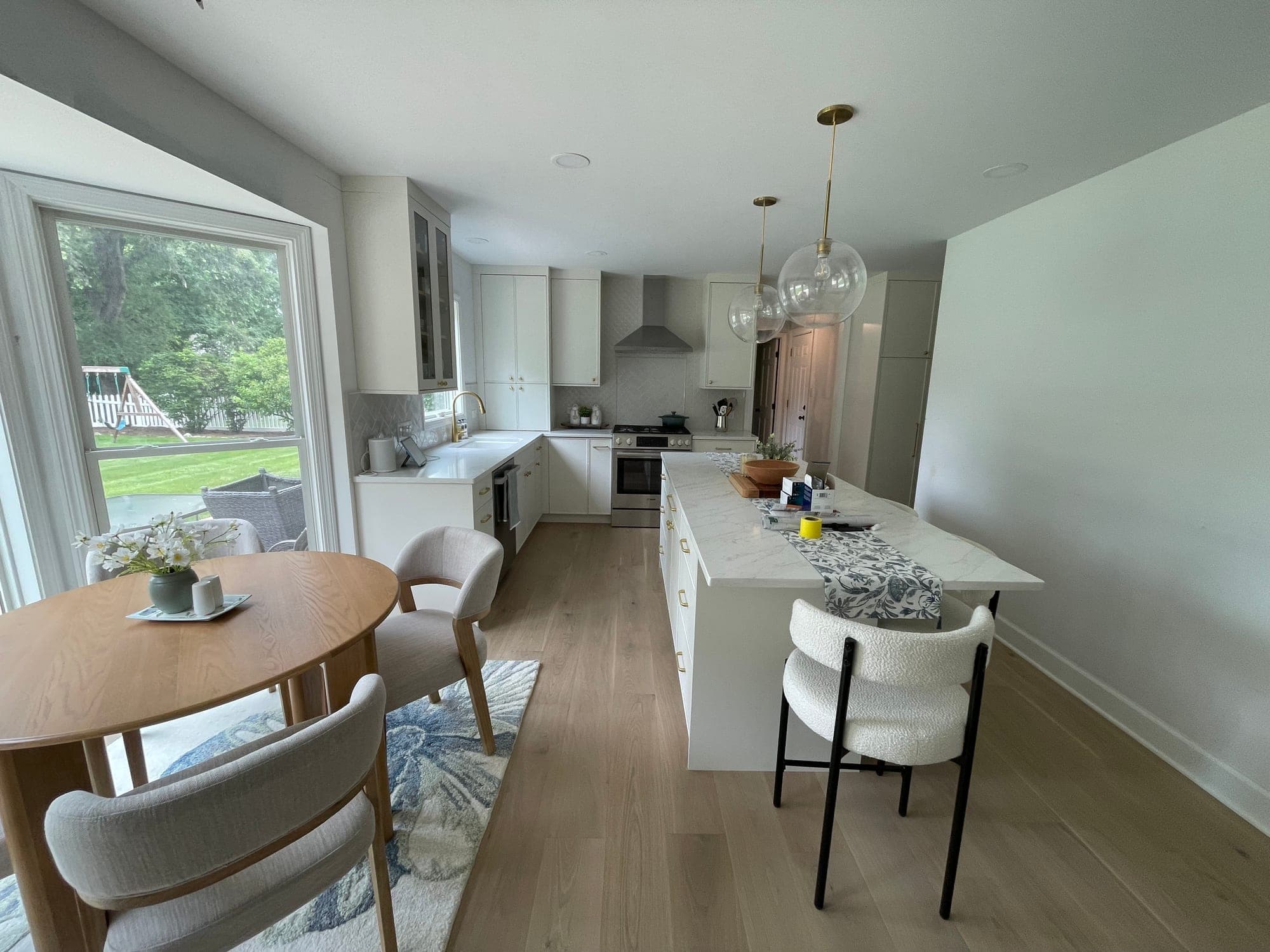 Long white kitchen with an island and globe pendants, open to a breakfast table beside a bay window
