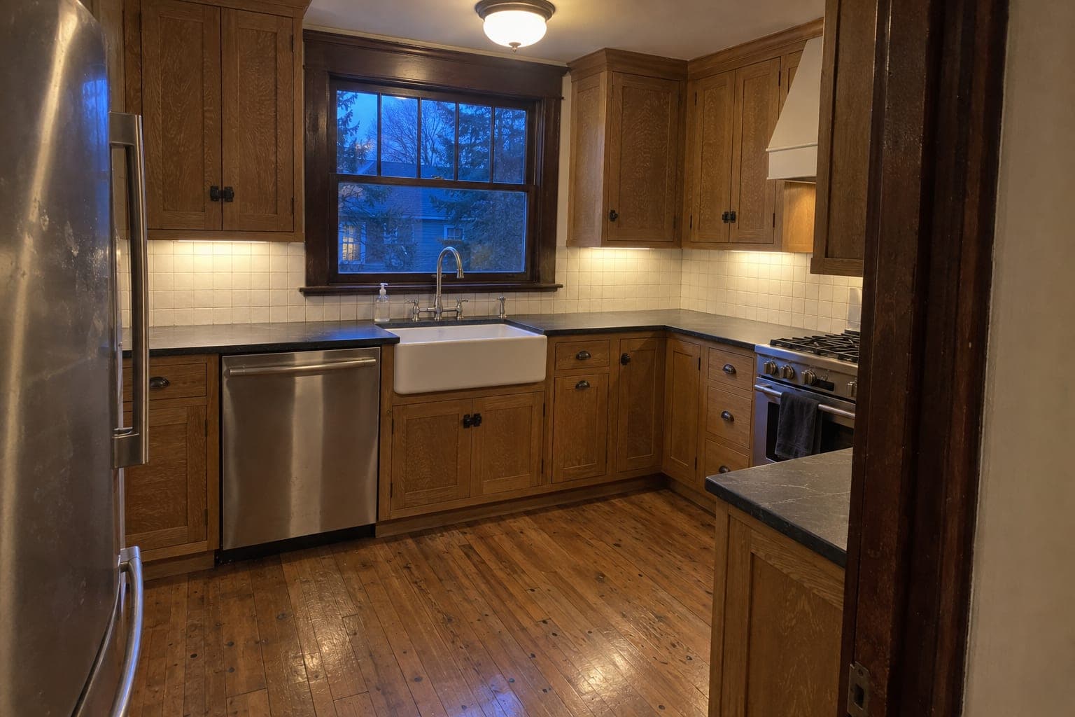 Craftsman style kitchen with quarter sawn oak cabinets, charcoal soapstone counters, a white apron sink, white square tile, dark original window trim and narrow oak flooring