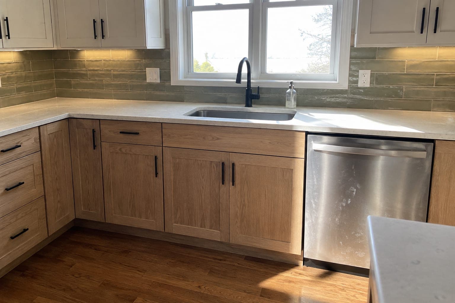 Sink wall with white oak lower cabinets, warm white uppers, quartz counters, olive ceramic tile, a black faucet, a stainless dishwasher and a refinished hardwood floor