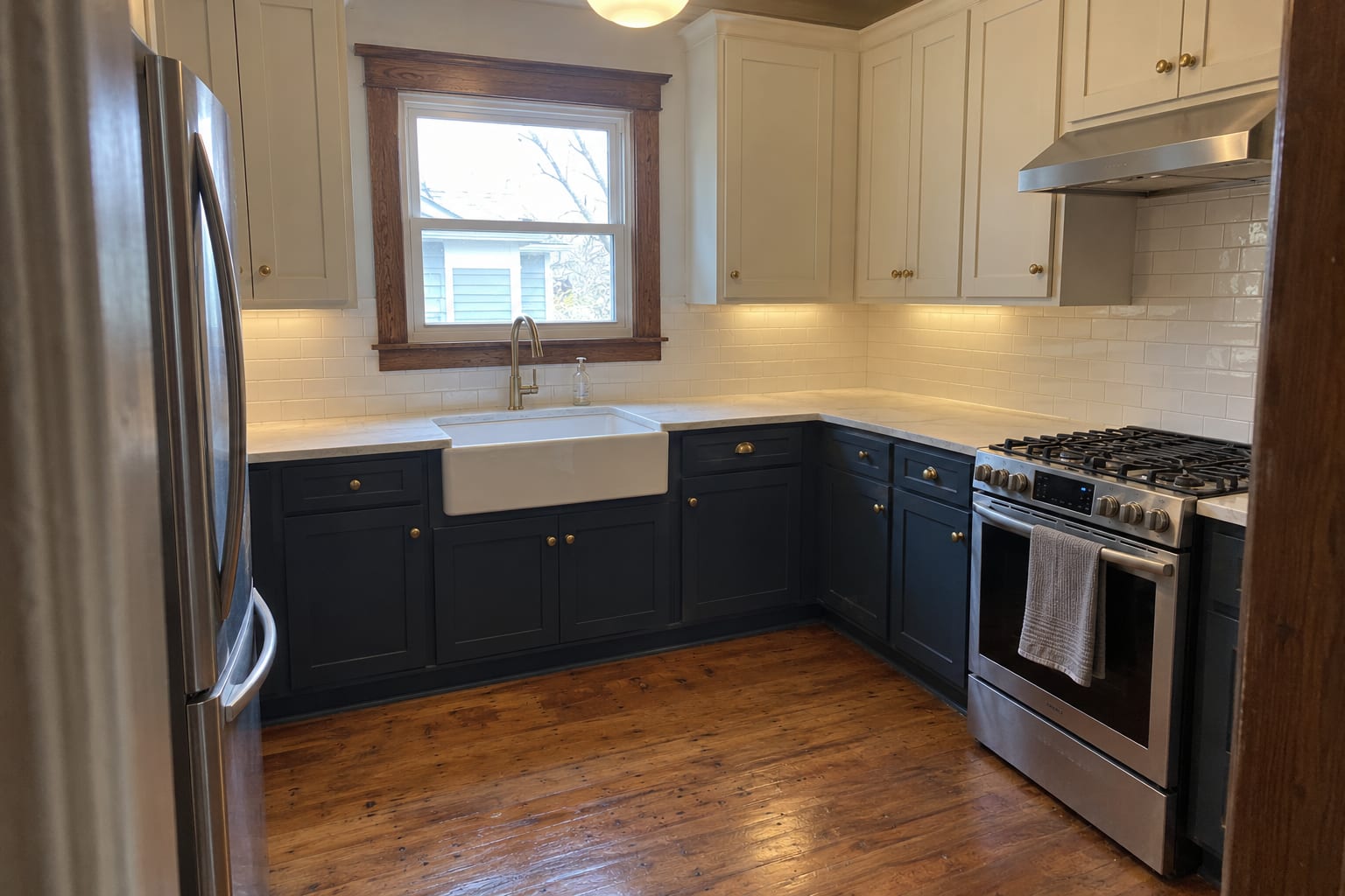 Small bungalow kitchen with cream upper cabinets, navy lower cabinets, a white apron sink, subway tile, original wood window trim and a restored oak floor