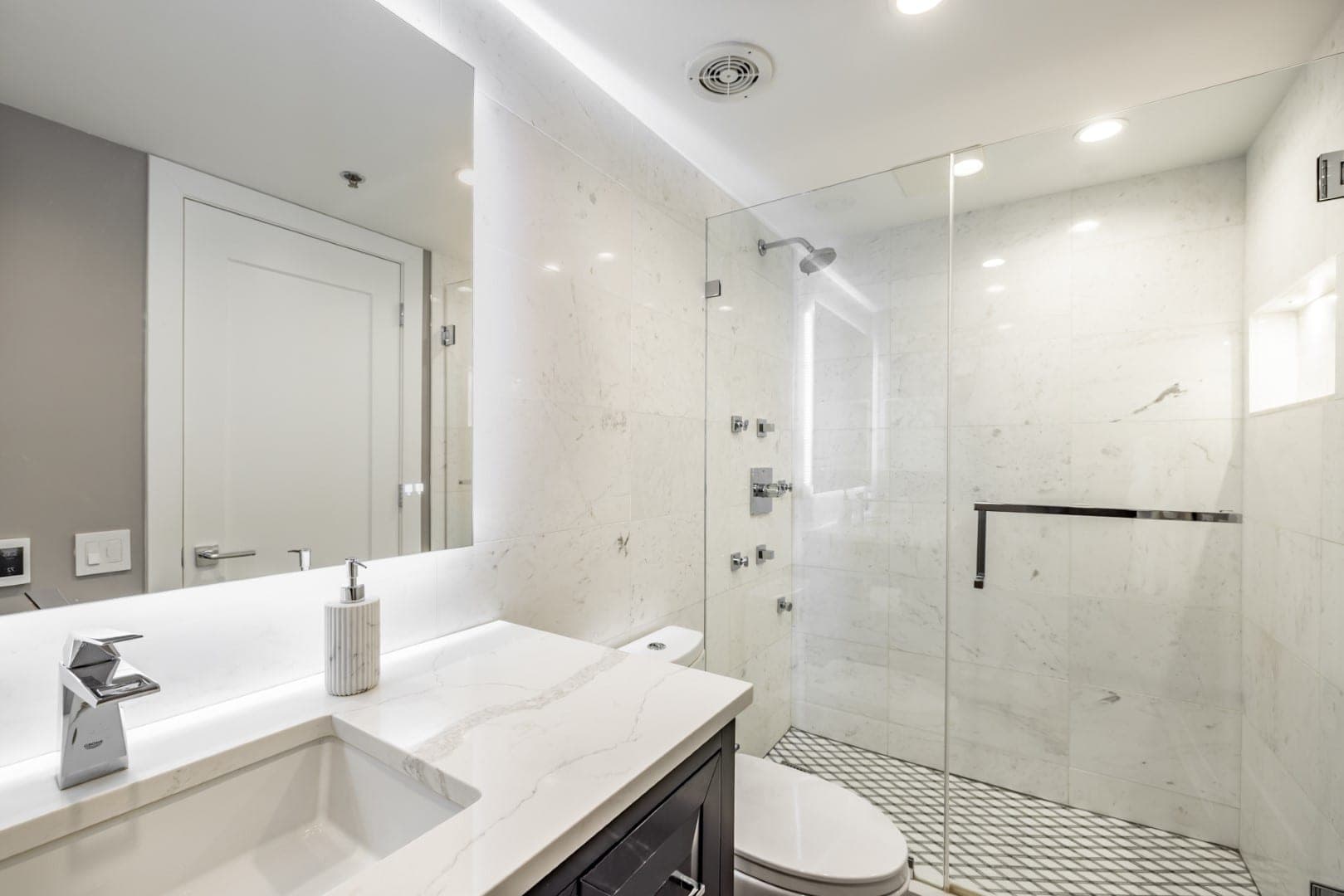Baño con regadera walk-in de vidrio en azulejo blanco veteado con chorros corporales y nicho, junto a un espejo iluminado