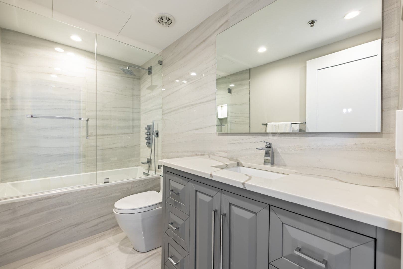 Baño con muros de losa veteada, tina con cancel de vidrio y mueble de lavabo gris con cubierta blanca y espejo sin marco