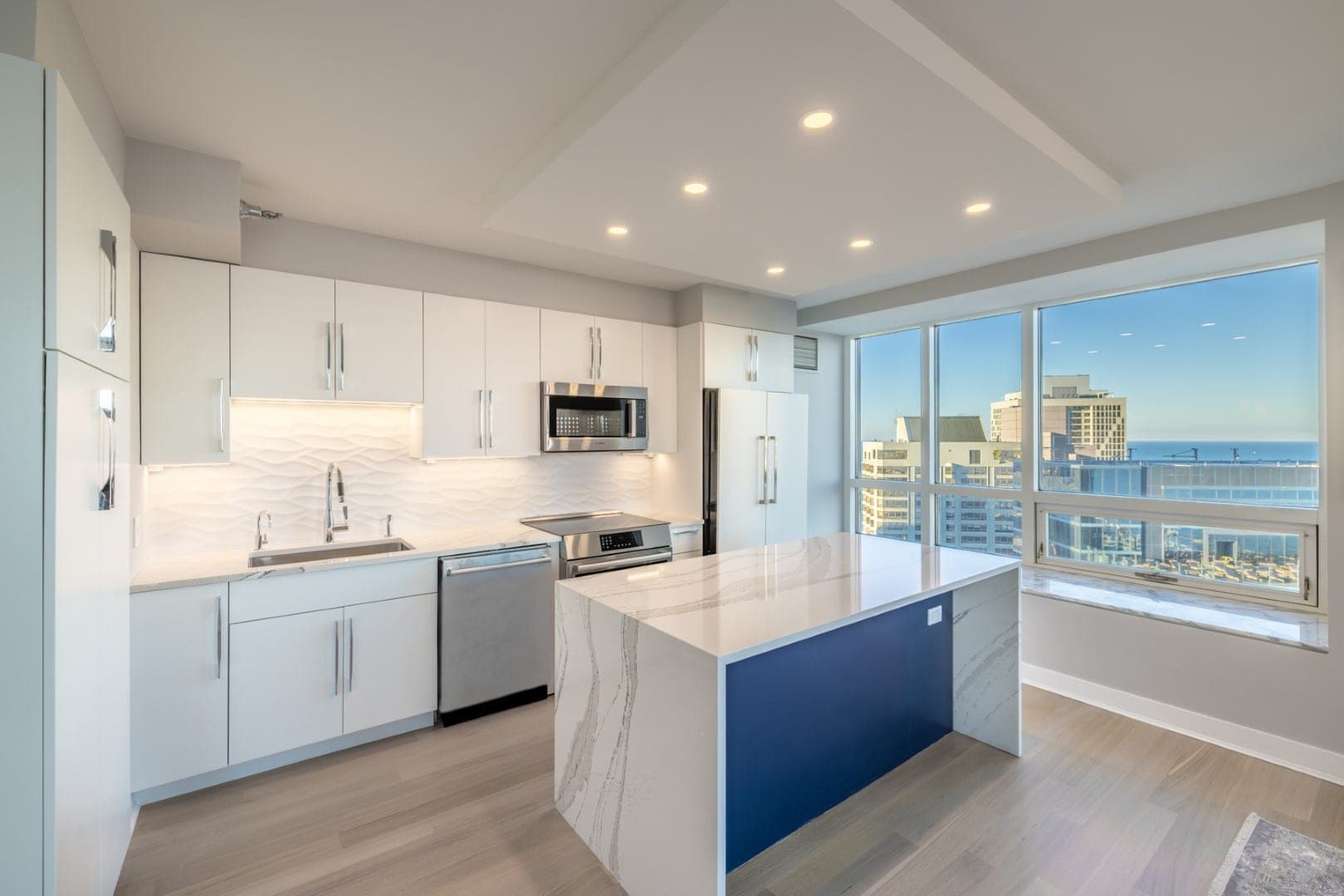 Cocina de condominio en un edificio alto, con gabinetes blancos lisos, isla azul marino en cascada, azulejo ondulado y vista al lago